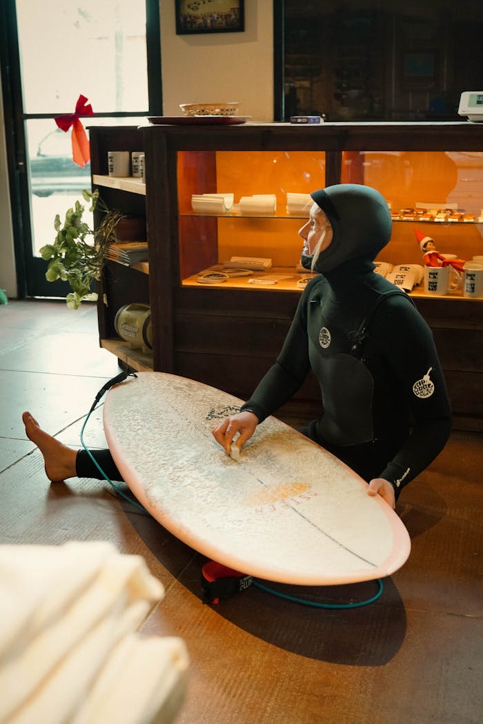 A female surfer in a wetsuit sitting indoors, waxing her surfboard.