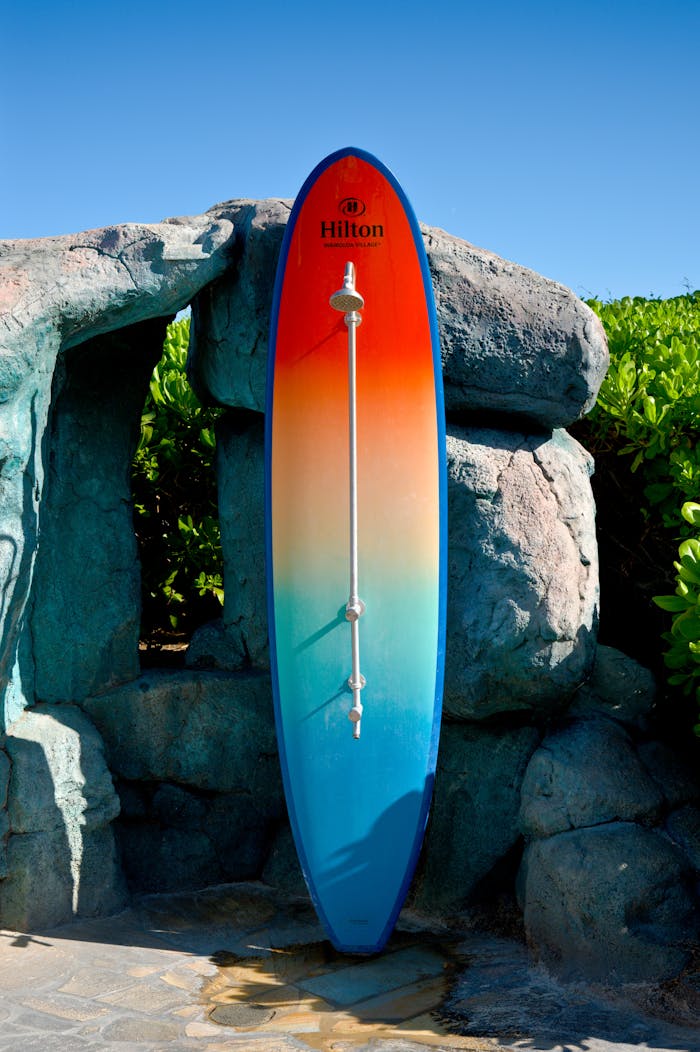Vibrant gradient surfboard leaning against rocks outdoors in daylight, Hilton logo visible.
